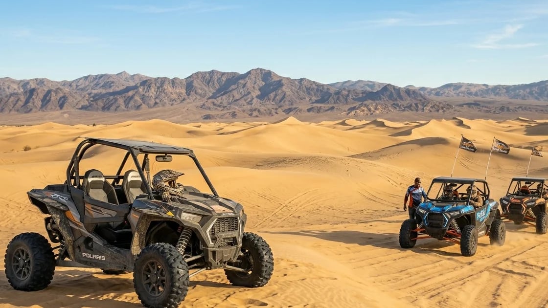 Vehículos todoterreno estacionados en un desierto arenoso con dunas doradas y montañas al fondo bajo un cielo azul claro