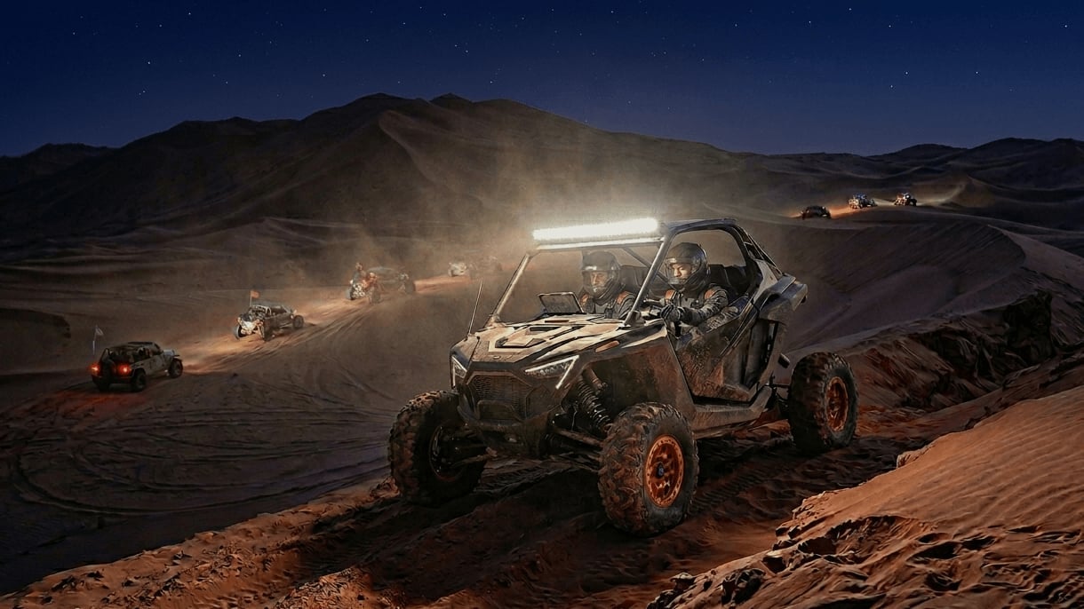 Off-road buggy with bright headlights racing across desert sand dunes at night with mountains in background